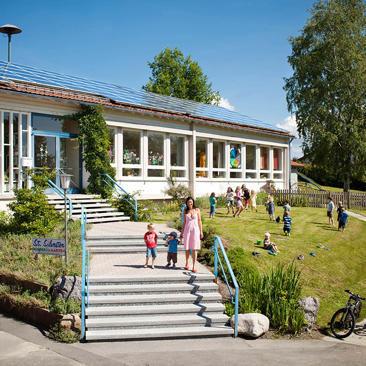 A female teacher with children in front of a kindergarten with a rooftop solar installation using premium REC solar panels