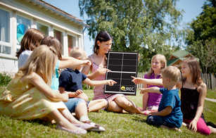 A group of children sitting on the grass point to an REC Alpha solar panel held by their teacher